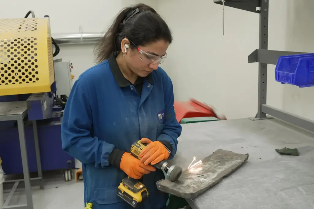 A worker uses a drill with a metal brush attachment to polish an ingot.