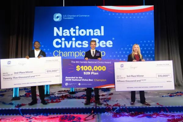 Three young participants on a stage at the National Civics Bee Championship hold large ceremonial checks for their placements, with the central winner receiving $100,000.